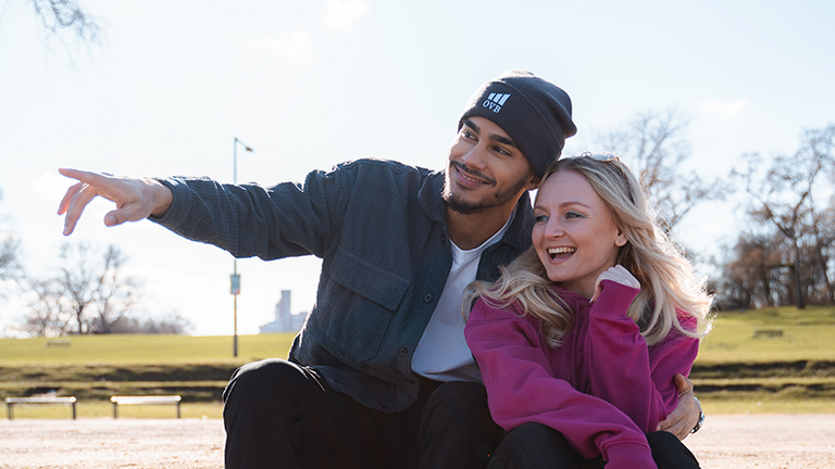 Mladi par na klupi u parku, smiju se zajedno – simbol cjeloživotnog financijskog savjetovanja od OVB-a.